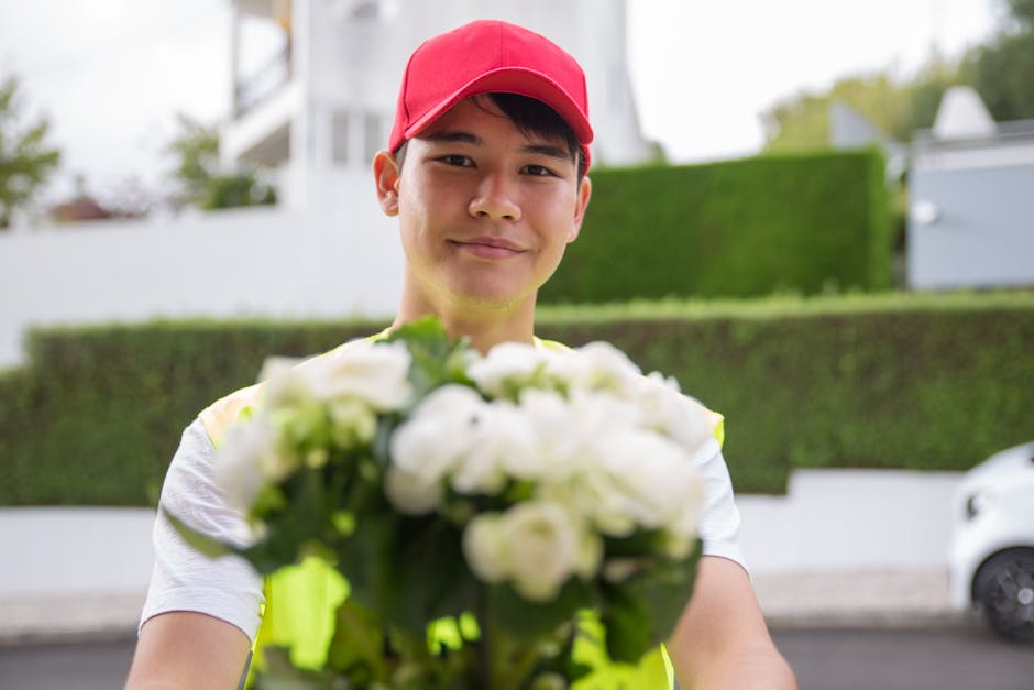 A delivery person wearing a red cap and a high-visibility vest is holding a bouquet of white flowers with green foliage towards the camera. The bouquet features fresh, closely packed petals and stems,