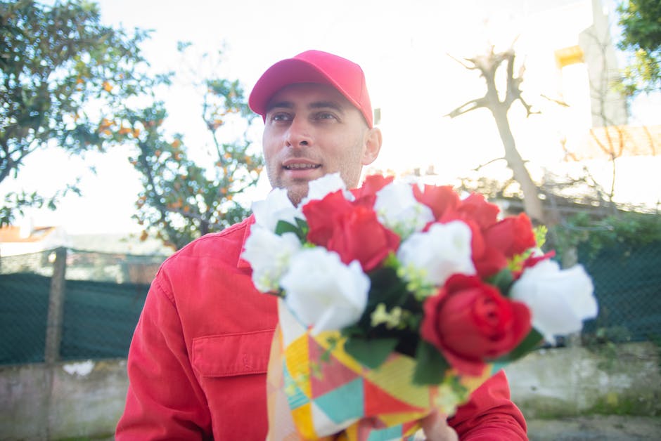 A male flower delivery courier from flowerdeliveryleytonstone.co.uk is seen outdoors, holding a vibrant bouquet of fresh flowers wrapped in decorative paper. The bouquet features red and white blooms,
