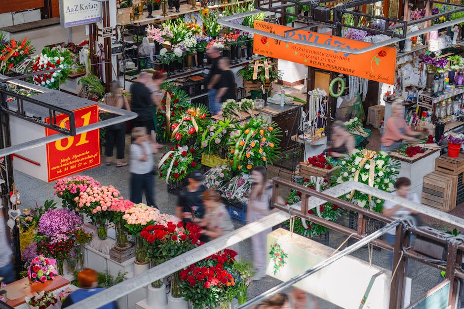 A busy indoor flower market featuring a wide variety of floral arrangements and bouquets. In the foreground, there are vibrant pink, red, and purple chrysanthemums and roses displayed in vases and pot